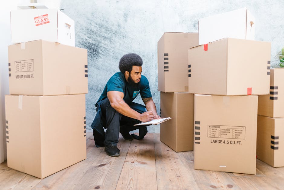 A man with dark skin and curly black hair, dressed in a teal shirt and black trousers, is crouching on the wooden floor inside a property during a home relocation process. He is holding a clipboard in