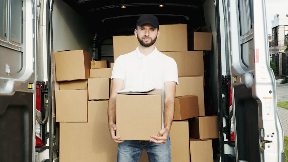 A man wearing a black cap, white shirt, and jeans stands inside the open back of a moving van, holding a cardboard box. The van is filled with various packed cardboard boxes of different sizes, stacked neatly to maximise space for a home relocation. The interior of the van shows a wooden floor and metal walls, with some boxes placed on the floor and others stacked against the sides. Outside, a driveway or street scene is partially visible, with part of a residential building and some greenery in the background, indicating an urban or suburban setting. The lighting is natural, suggesting daytime. The scene captures the loading process involved in furniture transport and packing during a professional removals service, as provided by Man and Van Ilford, emphasizing careful handling and efficient packing for house moves.