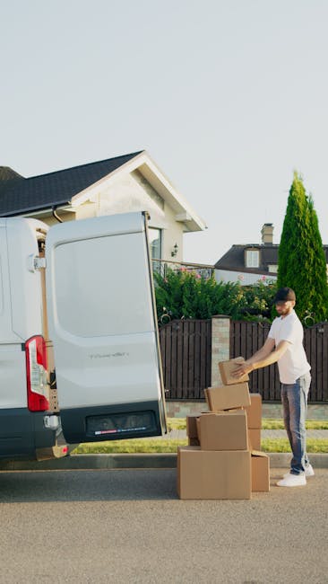 A man standing on a residential street beside a large cargo van with its rear door open, engaged in loading or unloading cardboard boxes during a home relocation. The man, dressed casually in a white t-shirt and jeans, is handling a box while three more boxes are stacked nearby on the pavement. The van is parked adjacent to a driveway, with a suburban house featuring a gabled roof, white walls, and a chimney visible in the background. Green trees and shrubs border the property, and the scene is illuminated by natural daylight, indicating clear weather. The image captures the manual loading process typical of furniture transport and packing and moving activities, with Man and Van Ilford occasionally mentioned in relation to efficient house removals and professional moving logistics.