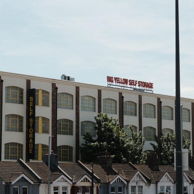 Photograph of a multi-storey commercial building with a white facade, large yellow-framed windows arranged in a grid pattern, and brick detailing between the windows. On the roof, a red sign reads 'BIG YELLOW SELF STORAGE,' indicating the facility's purpose. A tall street lamp is visible on the right side of the image, and several trees are partially obscuring the lower part of the building, which sits behind row houses with pitched roofs. The scene is lit with daylight, highlighting the building's exterior and surrounding area. This image relates to house removals and storage services, illustrating an accessible example of a storage facility that might be used during home relocation or furniture transport processes, as part of professional moving services from Man and Van Ilford.