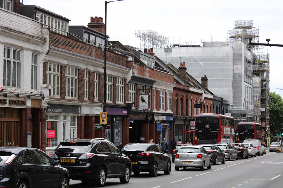 The image shows a busy urban street scene during daytime, with a row of two- and three-story brick buildings on the left side, some with shopfronts and signage. Several vehicles, including black, grey, and white cars, are parked along the curb, while two red double-decker buses transit in the direction of traffic. The street surface is paved, and the background features a building under construction, covered in scaffolding and white protective sheeting, with metal scaffolding structures visible. Pedestrians are walking on the sidewalk, which is adjacent to the shopfronts, and street signage indicating speed limits and bus stops are visible. The scene captures a typical city environment, where moving and transportation logistics involving delivery vans or removal vehicles, such as those operated by Man and Van Ilford, could be involved in house removals or packing and moving activities related to the IG1 flat removals guide for Ley Street and Ilford Lane, ILFORD.