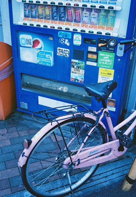 A pink bicycle with a black rear rack is parked in front of a blue vending machine on a pavement. The bicycle has a step-through frame, a black saddle, and a chain guard, with its front wheel turned slightly to the right. The vending machine features various drinks in plastic bottles and cans, with a coin slot, digital display, and several stickers and labels on its front. The area is outdoors, with a wooden planter on the left and a metal pole on the right, suggesting a street scene that could be relevant for house removals or moving logistics, such as parking and access considerations in urban environments, as seen in the Ilford removals access problems article at manandvanilford.com.