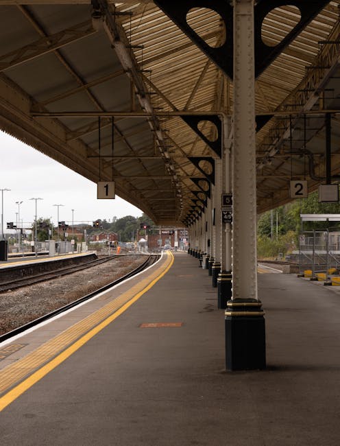 A view of Ilford train station platform with a canopy overhead supported by metal columns and beams, which are painted white with black bases. The platform is paved with dark asphalt and features yellow tactile paving along the edge nearest the trains. Numbered signs '1' and '2' are attached to the canopy structure, indicating different platform sections. In the background, train tracks curve away from the platform toward additional platforms, and distant buildings and trees are visible beyond the station. The station appears quiet with no passengers present in this moment. The setting illustrates a typical outdoor railway station environment as part of a home relocation or moving logistics process, highlighting the importance of reliable transport routes, as covered on the page about Ilford station removals and best local moving routes at manandvanilford.com.
