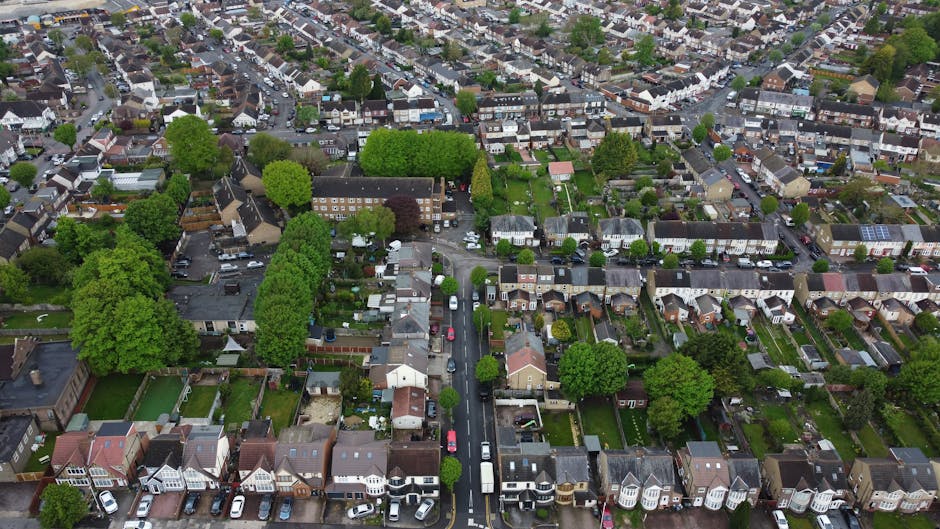 Aerial view of a suburban residential area with rows of terraced and detached houses, many with small gardens and driveways, surrounded by a variety of lush green trees and well-maintained yards. The street in the foreground features parked cars along both sides, with a white van and other vehicles visible, indicating active house removals or moving preparations. The image captures the quiet neighborhood environment during daytime, showcasing the density of housing typical for house relocations and local moving services in the ILFORD area. The lighting is natural, highlighting the different roof types, garden layouts, and tree canopies, providing a comprehensive view relevant to home relocation logistics, packing, and furniture transport supported by companies like Man and Van Ilford.