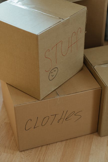 Close-up image of three brown cardboard moving boxes stacked indoors on a wooden floor, with handwritten labels indicating their contents. The top box is labeled 'STUFF' in red marker, accompanied by a simple smiley face drawn beneath the word. The middle box is labeled 'CLOTHES' in black marker. The boxes are sealed with packing tape and appear to be part of a home relocation or packing process, typical of furniture transport and moving services. The background reveals a partially visible doorway and additional packaging materials, suggesting an active packing or unloading area as part of a house removals operation managed by Man and Van Ilford. The lighting is natural, highlighting the secure stacking and clear labeling of the boxes, supporting efficient moving logistics and packing organization during an urgent or same-day move.