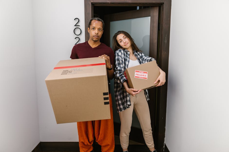 A man and a woman standing inside a residential property near a doorway, both holding cardboard boxes during a home relocation. The man, wearing a maroon t-shirt and orange trousers, holds a large box with red packing tape and labels. The woman, dressed in a checked shirt over a white top and beige trousers, holds a smaller box marked with a red 'Fragile' sticker. The background shows a plain white wall with the house number to the left and a wooden doorframe, indicating they are preparing for a furniture transport or loading process as part of a same-day removal service. The lighting is natural, illuminating the interior, and the scene depicts the early stages of packing and moving logistics, characteristic of professional removals by Man and Van Ilford, related to house removals in the ILFORD area.
